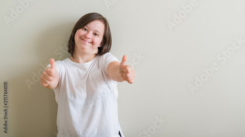 Down syndrome woman standing over wall approving doing positive gesture with hand, thumbs up smiling and happy for success. Looking at the camera, winner gesture. photo