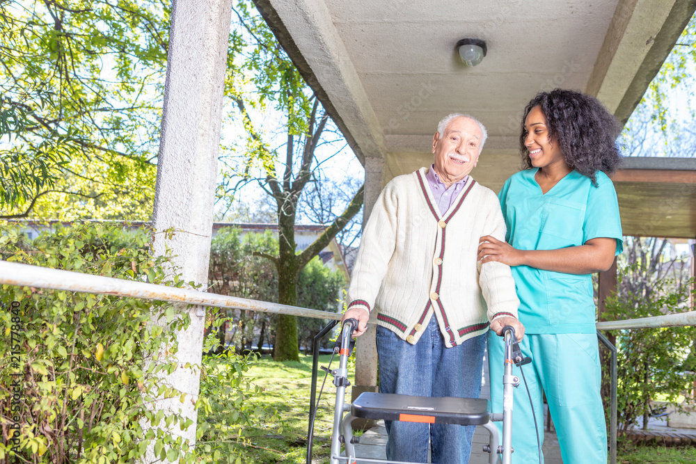 African nurse helping elderly man with walker in rehab facility hallway
