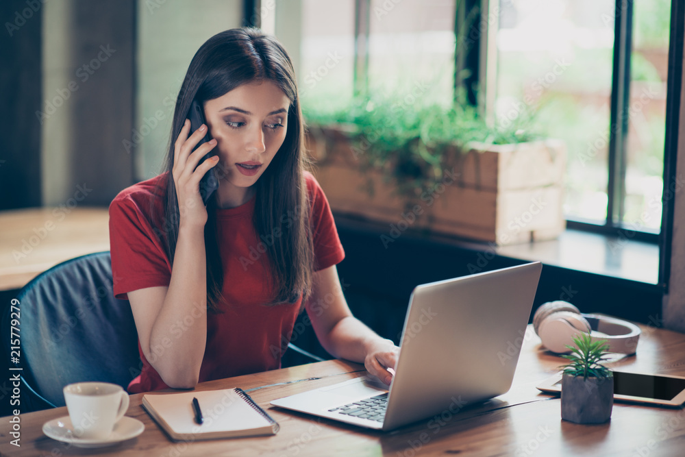 Freelancer business girl in red polo working issues by phone, looks at laptop and fixes errors in documents sitting in cafe Freelancer business girl in red polo working issues by phone, looks at laptop and fixes errors in documents sitting in cafe