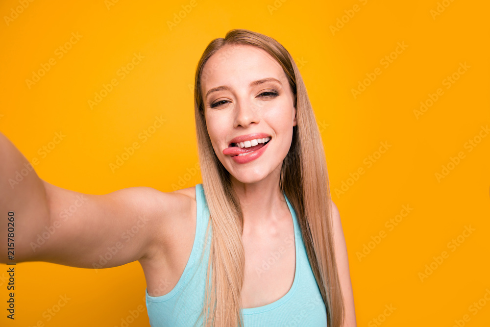 Portrait of happy foolish girl looking camera with tongue out and makes selfie on the front camera of her smartphone isolated on shine yellow background Portrait of happy foolish girl looking camera with tongue out and makes selfie on the front camera of her smartphone isolated on shine yellow background