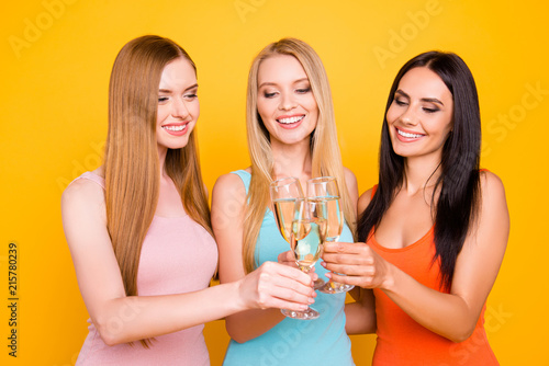 Portrait of cheerful positive girls holding alcohol beverage in hands clicking glasses, blonde brunette ginger enjoying free time together having beaming smiles isolated on bright yellow background