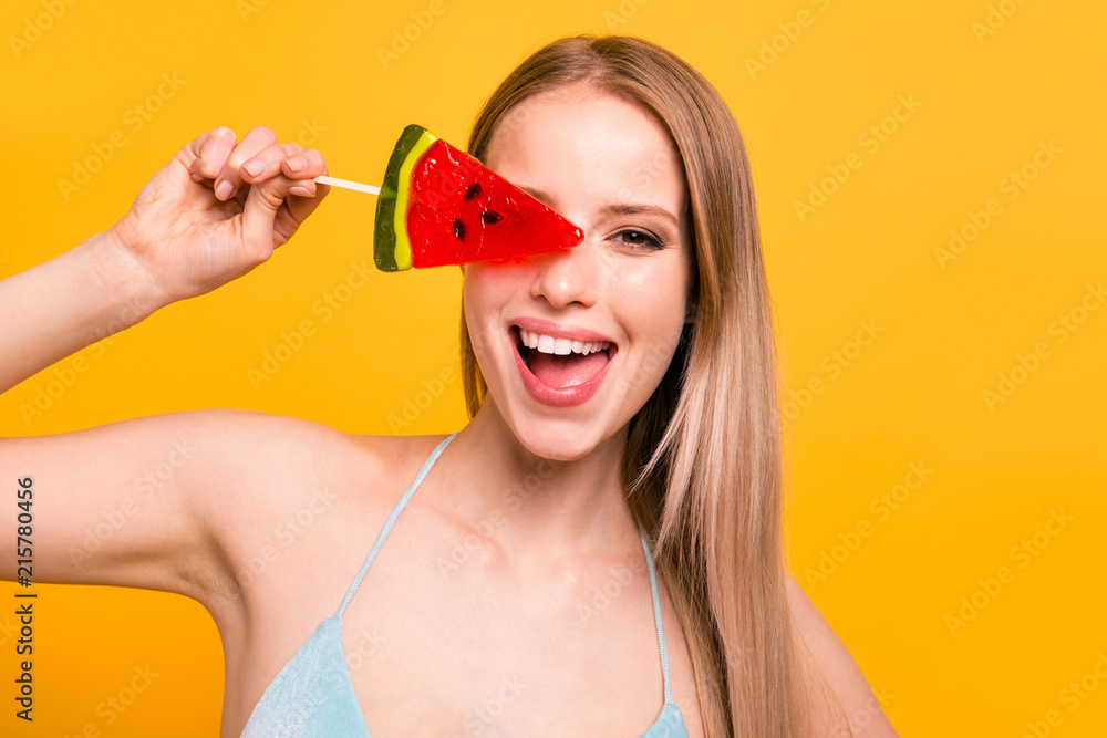 Portrait of attractive nice cute straight-haired smiling blonde girl, wearing blue swimsuit, fooling, hiding one eye behind candy on stick. Isolated over yellow background