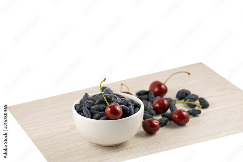 Fresh ripe honeysuckle berries and cherry in a ceramic bowl on a wooden board. Healthy breakfast. White bowl of honeysuckle. Fresh ripe honeysuckle berries and cherry in a ceramic bowl on a wooden board. Healthy breakfast. White bowl of honeysuckle.