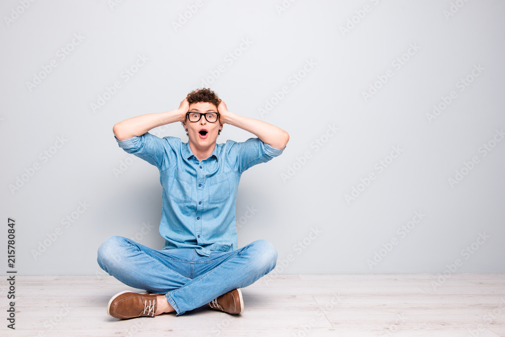 Handsome curly-haired amazed young guy wearing casual jeans denim, shoes and glasses, sitting on floor with crossed legs, wow gesture, facial expression. Copy space. Isolated on light grey background