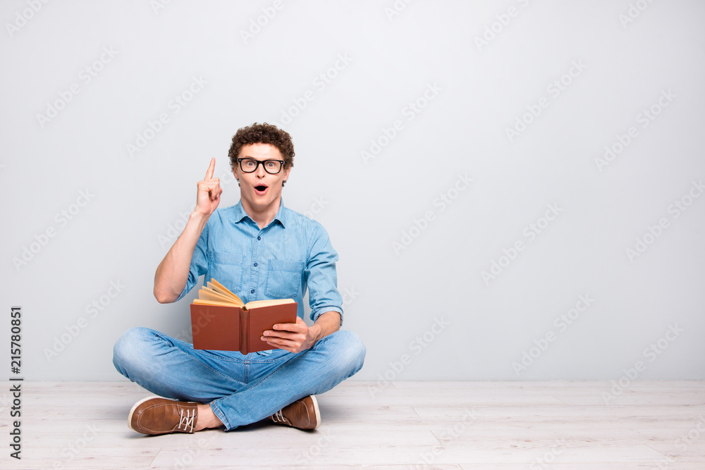 Good thought! Handsome curly-haired amazed guy wearing casual jeans denim, shoes, glasses, sitting on floor with crossed legs, reading book, pointing finger up. Copy space. Isolated on grey background Good thought! Handsome curly-haired amazed guy wearing casual jeans denim, shoes, glasses, sitting on floor with crossed legs, reading book, pointing finger up. Copy space. Isolated on grey background