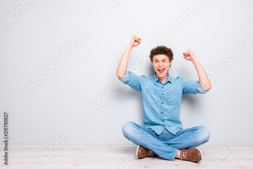 Handsome curly-haired cheerful young guy wearing casual jeans denim, shoes, sitting on floor with crossed legs, raising hands up. Copy space. Isolated over light grey background