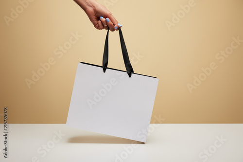 cropped image of woman holding white shopping bag above white table isolated on beige