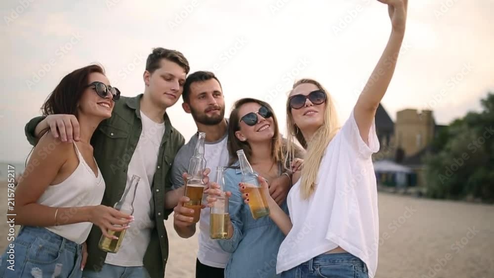 Group of friends having fun enjoying a beverage and relaxing on the beach at sunset in slow motion. Young men and women drink beer standing on a sand in the warm summer evening. Group of friends having fun enjoying a beverage and relaxing on the beach at sunset in slow motion. Young men and women drink beer standing on a sand in the warm summer evening.