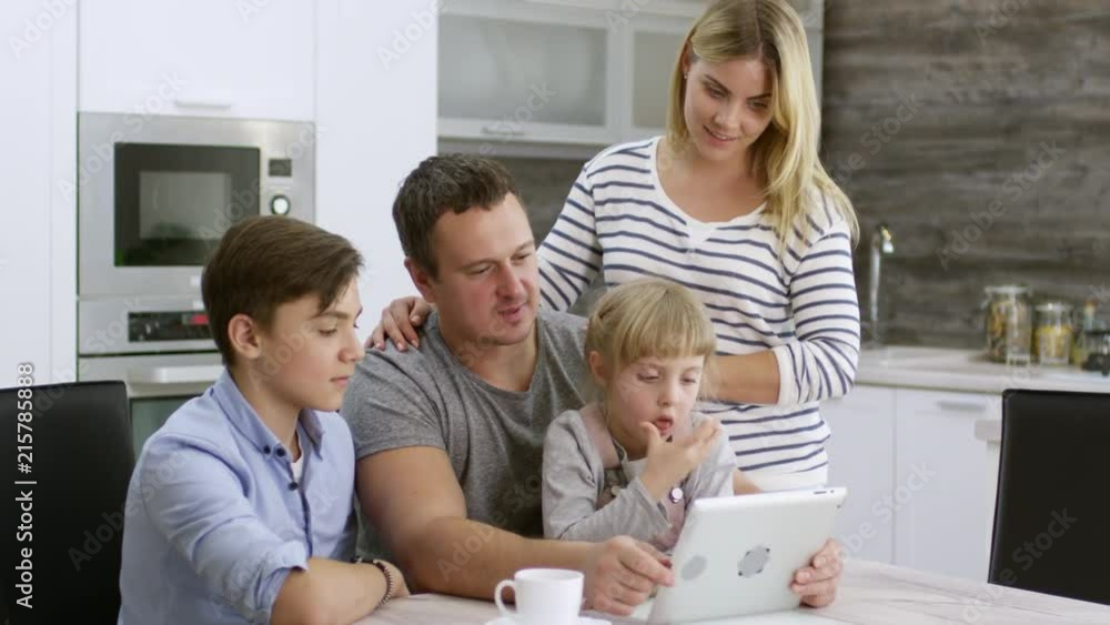 Beautiful mother, father, little daughter and teenage son watching something on digital tablet and talking at kitchen table Beautiful mother, father, little daughter and teenage son watching something on digital tablet and talking at kitchen table