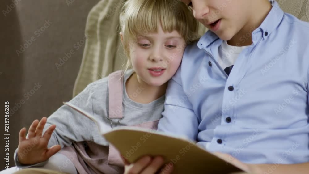 Young boy sitting with pretty little sister on sofa in the living room and reading fairytale to her Young boy sitting with pretty little sister on sofa in the living room and reading fairytale to her