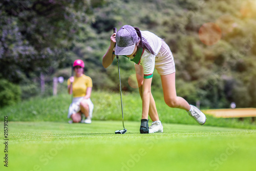 young woman golf player taking a golf ball in the hole on the green, with golfmate or challenger competitor exciting or enforcement in background, exciting or enforcement between players