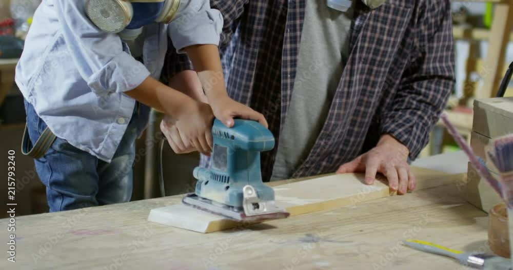 Tilt up of Asian father and little boy with respirators using belt sander on wood board in carpentry workshop Tilt up of Asian father and little boy with respirators using belt sander on wood board in carpentry workshop
