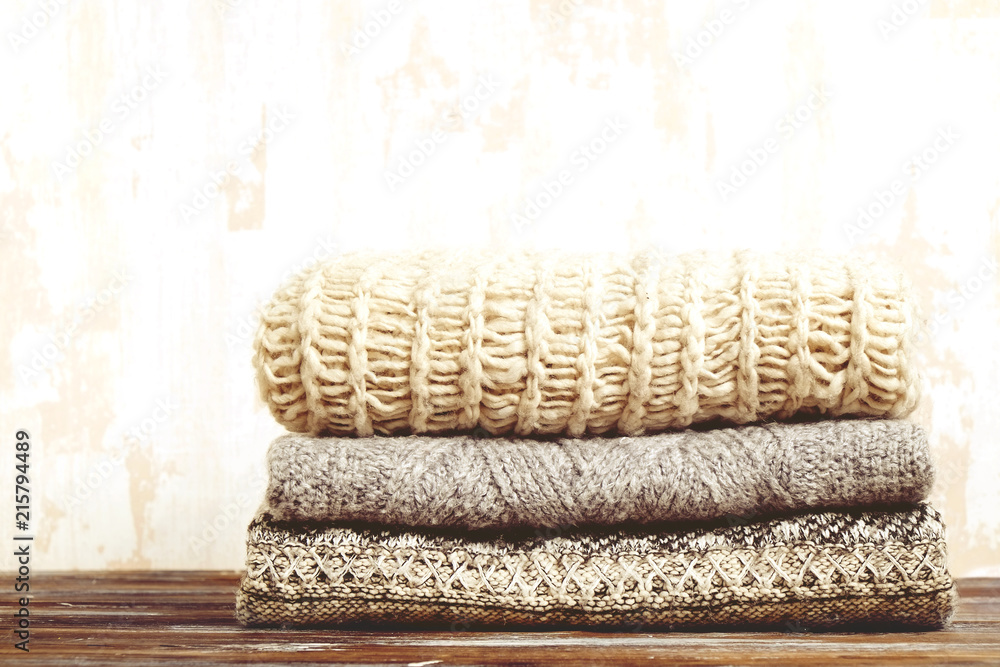 Bunch of knitted warm pastel color sweaters with different knitting patterns folded in stack on brown wooden table, grunged texture wall background. Fall winter season knitwear. Close up, copy space