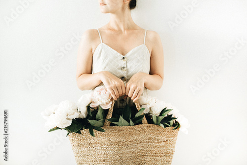 Fototapeta Naklejka Na Ścianę i Meble -  Young pretty woman holding straw bag with white peony flowers on white background. Summer concept.