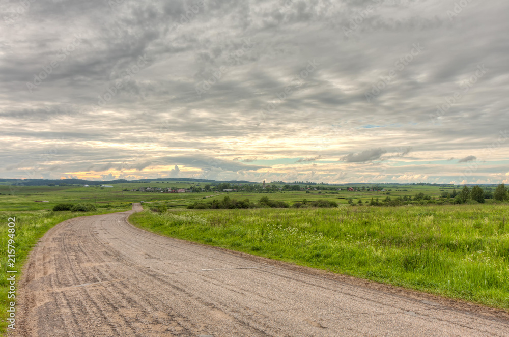 Vanishing dirt road to village with church against cloudy sky background. Derevni, Yaroslavsky region, Russia. 