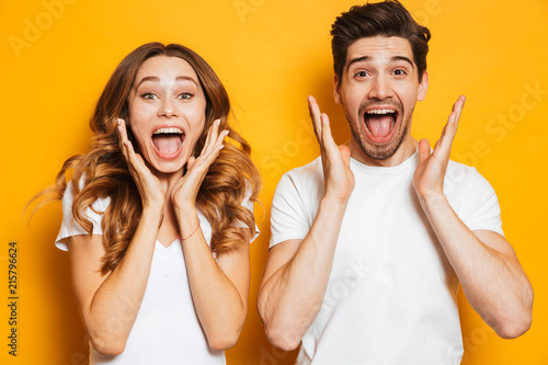 Photo of emotional couple man and woman in basic clothing screaming in surprise or delight and raising arms, isolated over yellow background