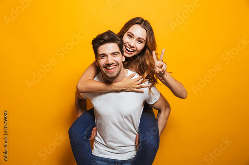 Image of caucasian couple having fun while man piggybacking joyful woman, isolated over yellow background