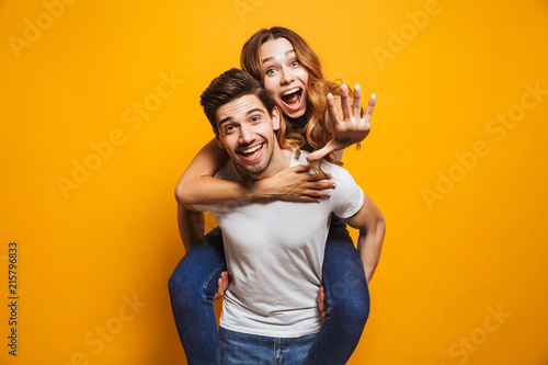 Photo of caucasian couple smiling while man piggybacking excited woman, isolated over yellow background