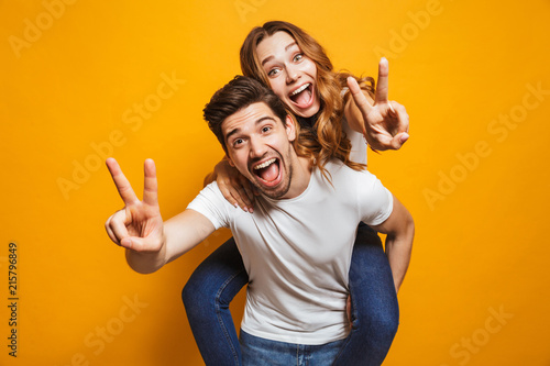 Image of caucasian man having fun and giving piggyback ride to joyful woman, isolated over yellow background