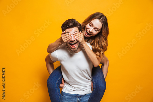 Image of loving couple having fun while man piggybacking joyful woman, isolated over yellow background