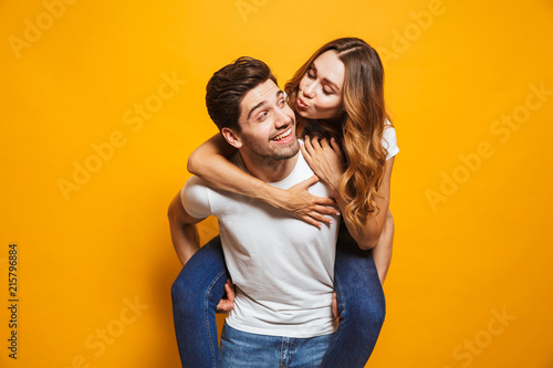 Photo of happy couple having fun while man carrying beautiful woman on his back, isolated over yellow background
