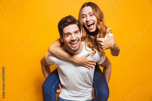 Image of excited couple having fun while man piggybacking joyful woman with thumb up, isolated over yellow background photo