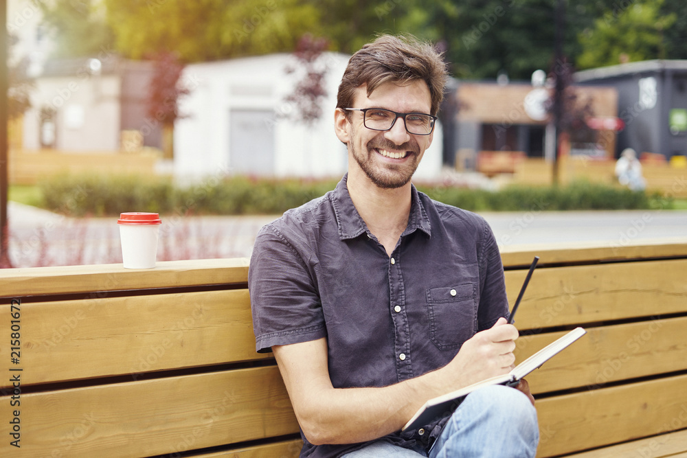 Obraz premium Handsome young man smiling face writing in a note book sitting outside. Guy wearing glasses alone working. Concept of education students youth.