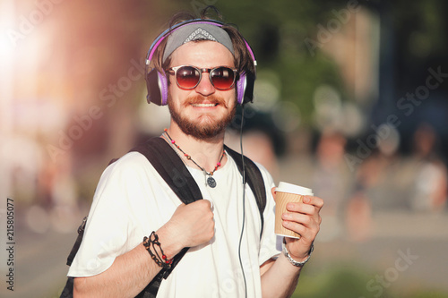 Handsome bearded hipster boy in sunglasses and headphones walking with backpack at the sunset on the street, he drinking coffee and listening music photo