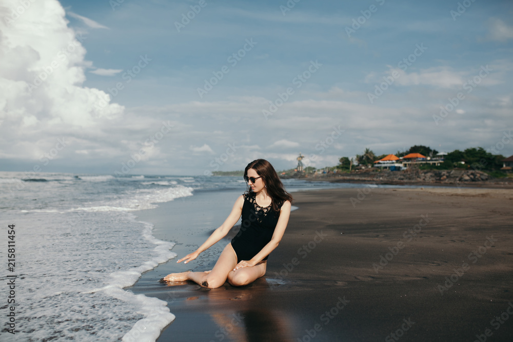 Happy smile brunette Woman playing with waves in black swimsuit and denim jacket on ocean background on black sand beach