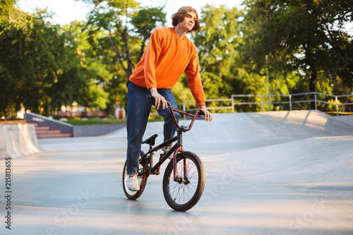 Young thoughtful guy in orange pullover and jeans dreamily riding bicycle at skatepark