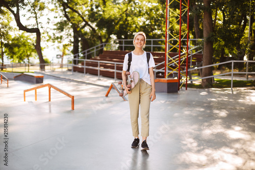 Beautiful smiling girl with skateboard in hand happily looking in camera with modern skatepark on background