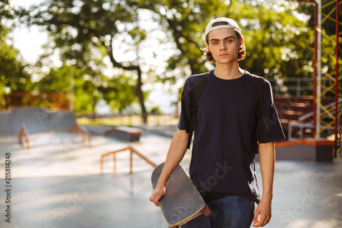 Young serious guy in black T-shirt and white cap thoughtfully looking in camera while holding skateboard in hand with modern skatepark on background