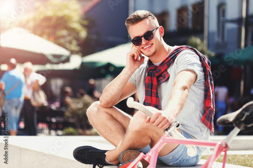 Handsome young hipster man in sunglasses sitting near the bicycle and speaking on the smartphone in the sunny street