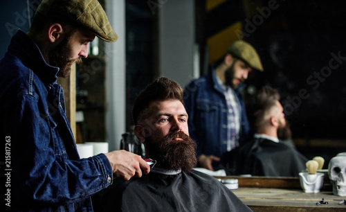 Hipster with beard covered with cape trimming by professional barber in stylish barbershop. Barber busy with grooming beard of hipster client, mirror reflexion on background. Grooming concept