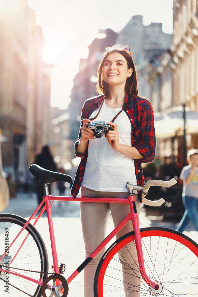 Obraz premium Portrait of young stylish hipster romantic long-hair girl in sunglasses staying with pink bicycle and making photos of architecture at sunny summer street