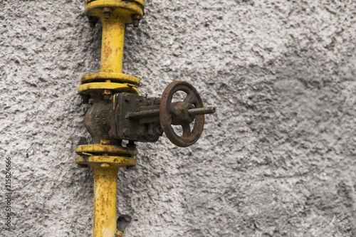 Old weathered gas crane and pipe on the background of a gray wall. Old gas gate of yellow colour is on a pipe and blocks a pipe. The pipeline with cranes for gas giving on the wall. photo