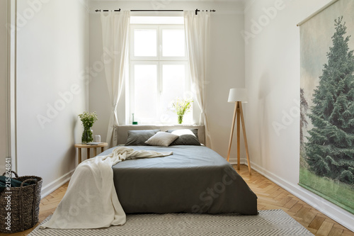 Blanket on black bed in bright bedroom interior with flowers, lamp and drapes at window. Real photo