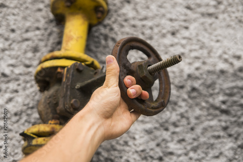 Man holding old weathered gas crane on the background of a gray wall. Old gas gate of yellow colour is on a pipe and blocks a pipe. The pipeline with cranes for gas giving on the wall. photo