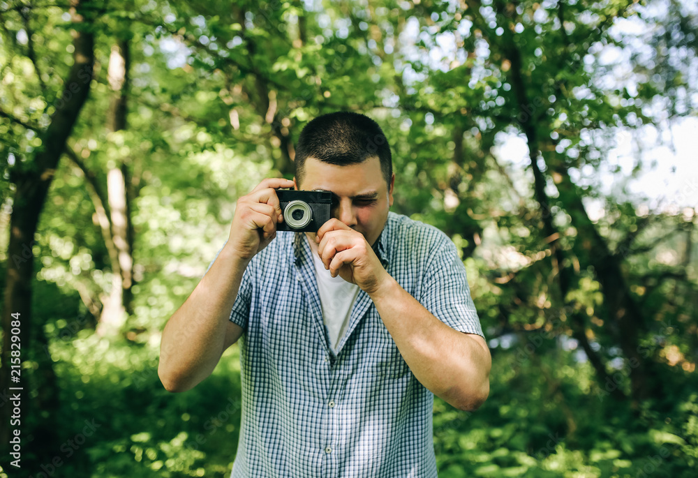 Young guy in checkered shirt is standing in the green forest with an old vintage photo camera. Hipster taking photos outdoors. Lifestyle travelling shot. Young guy in checkered shirt is standing in the green forest with an old vintage photo camera. Hipster taking photos outdoors. Lifestyle travelling shot.