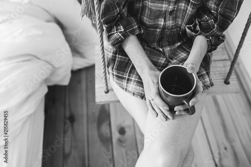 Beautiful girl is sitting on swing in loft minimalist interior. Young woman in men's checkered shirt is drinking coffee. Cozy morning photo.