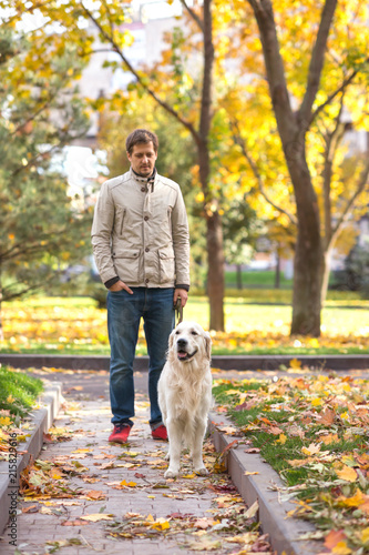 The young man wearing  jeans and a grey jacket and   white Labrador dog  are walking in the autumn park.