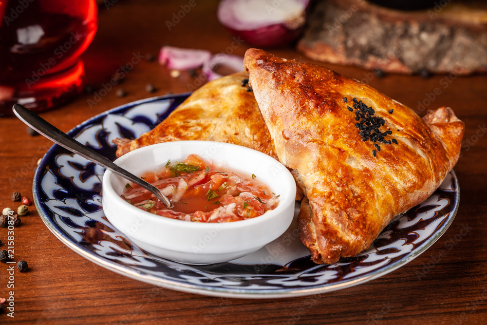 Uzbek samsa in national plate with tomato sauce on a wooden table. end of the eastern baking. Uzbek samsa in national plate with tomato sauce on a wooden table. end of the eastern baking.