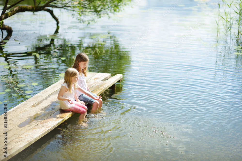 Fototapeta premium Two cute little girls sitting on a wooden platform by the river or lake dipping their feet in the water on warm summer day