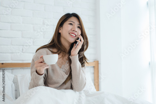 Asian woman using the smartphone on her bed while holding cup of coffee in the morning. Beautiful asian woman enjoying warm coffee and talking on telephone in her bedroom. lifestyle asia woman concept