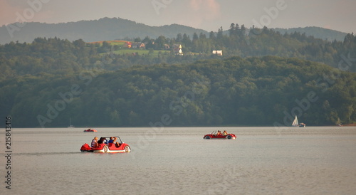 Jezioro Solińskie, Bieszczady, Polska, rowerki wodne z turystami na wodzie, wzgózra porośnięte gęstym lasem na przeciwnym brzegu akwenu, na jednym ze wzgórz zabudowania, letnia atmosfera