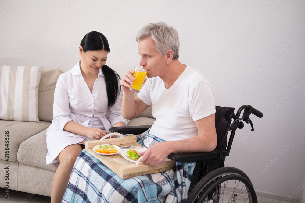 Beautiful asian nurse with her hair in sleek ponytail sitting on couch next to male patient with gray hair in wheelchair eating lunch. Beautiful asian nurse with her hair in sleek ponytail sitting on couch next to male patient with gray hair in wheelchair eating lunch.