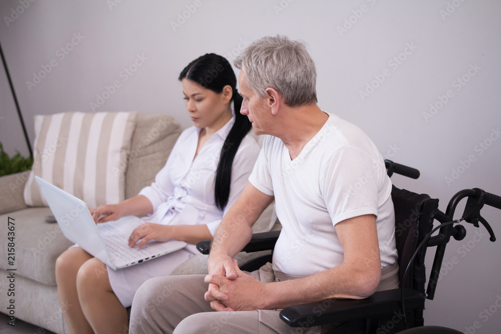 Nurse typing on computer sitting next to disabled patient. Young beautiful caregiver showing aged man how laptop works while he sits in wheelchair watching her. Nurse typing on computer sitting next to disabled patient. Young beautiful caregiver showing aged man how laptop works while he sits in wheelchair watching her.