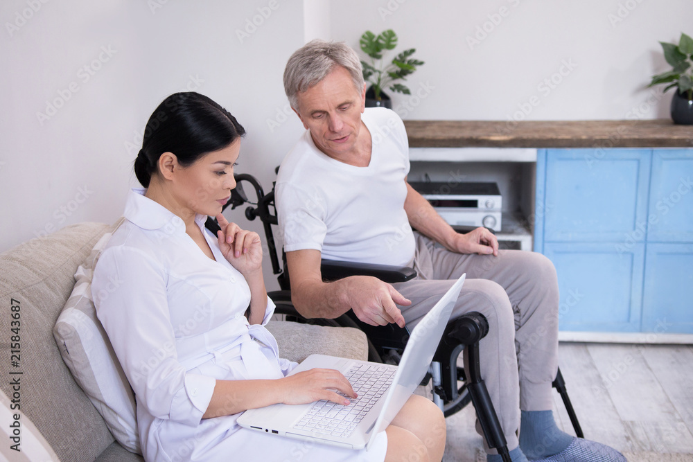Man in wheelchair pointing at laptop screen. Young asian nurse working on computer sitting next to aged disabled male patient who is showing her something on screen. Man in wheelchair pointing at laptop screen. Young asian nurse working on computer sitting next to aged disabled male patient who is showing her something on screen.