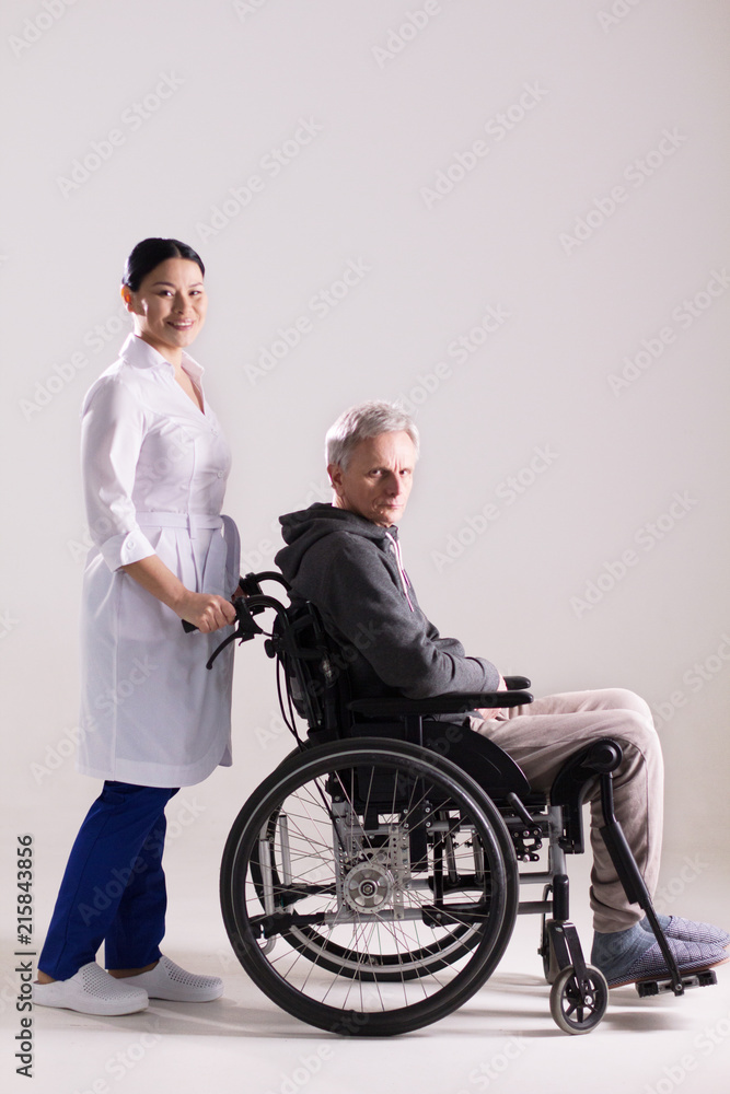 Full length of female nurse pushing wheelchair with man in it. Studio shot of beautiful asian woman working as doctor giving disbaled man on chair push. Full length of female nurse pushing wheelchair with man in it. Studio shot of beautiful asian woman working as doctor giving disbaled man on chair push.