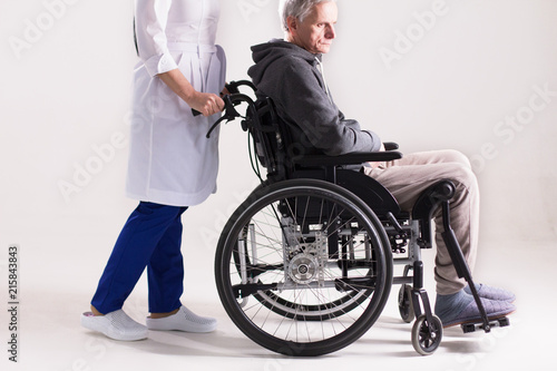 Studio shot of doctor pushing wheelchair with man it. Cropped picture of doctor in white coat and blue pants pushing wheelchair with elderly man sitting inside. © Svyatoslav Lypynskyy